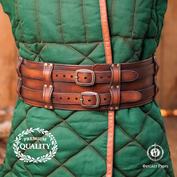 May include: A wide brown leather belt featuring two horizontal straps with metal buckles and rivets, worn over a green quilted garment. The belt has a distressed finish and is secured with two buckles. A logo with the text PREMIUM QUALITY is visible.
