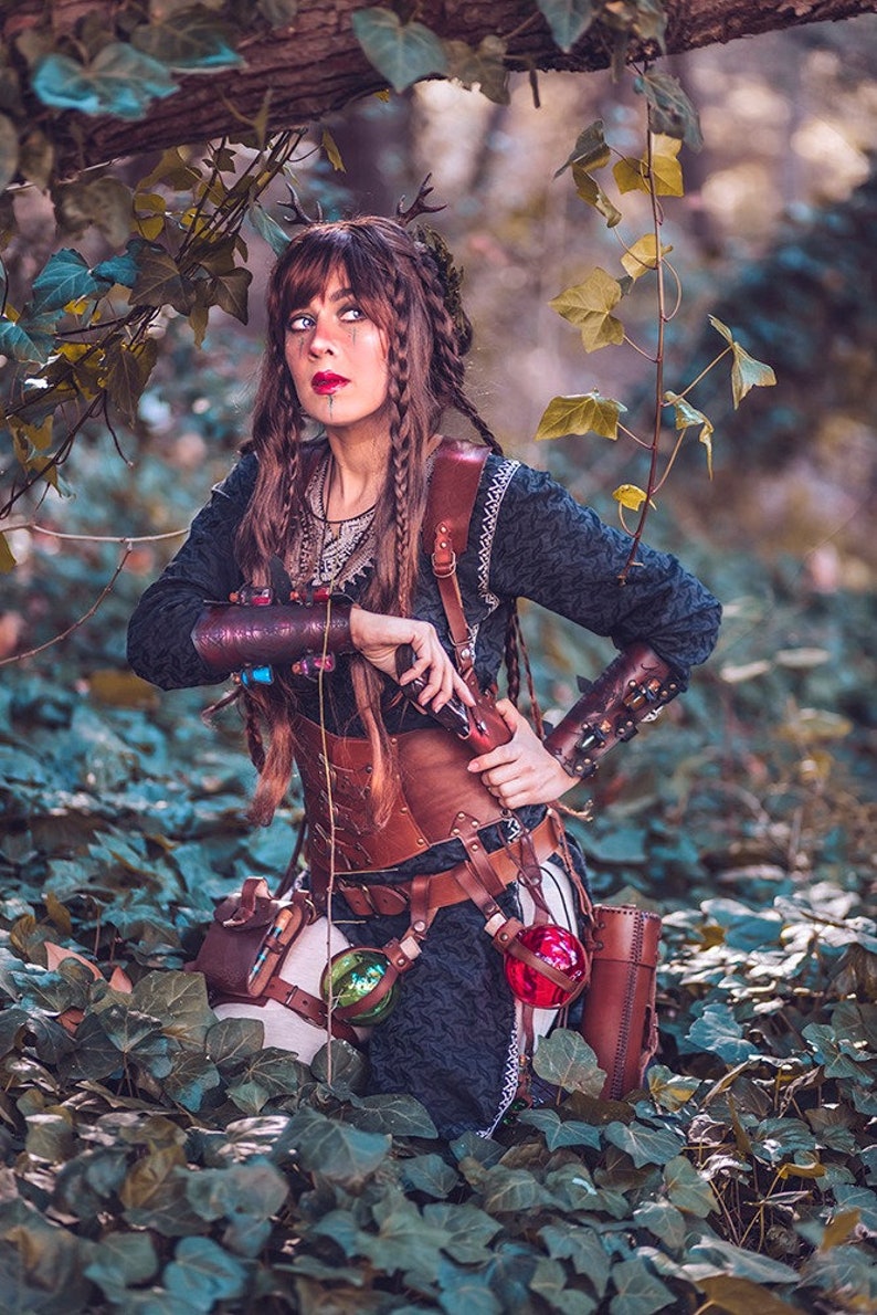 Female adventurer in the forest wearing the old brown leather corset model. The new edition is crafted in aged brown with nickel hardware and more elegant buckles. Designed for witches, alchemists, and fantasy LARP characters, this steampunk waist cincher combines historical style and eco-tanned leather craftsmanship, ideal for renaissance fairs, gothic events, and fantasy festivals.