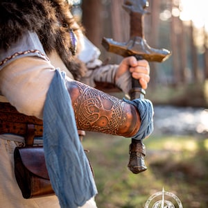 Close-up of a warrior gripping a sword with both hands, showcasing brown leather bracers with tree of life embossing. The outfit combines linen, fur, and blue wraps, ideal for larp barbarian gear, viking costume armor, leather vambrace handmade, or renaissance cosplay outfit. Forest lighting highlights the texture and aged finish of the embossed leatherwork. Yggdrasil Viking leather bracer world tree vambrace for larps cosplay yule high-quality full-grain vegetable tanned leather embossed armor
