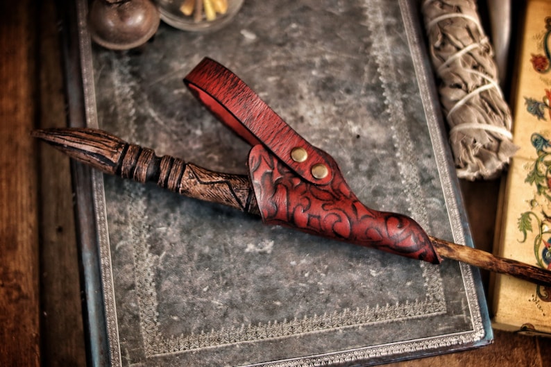 A red embossed leather wand holster rests on a weathered metal covered book while holding a carved wand. The holster features ornate patterns, brass toned rivets and a vivid hand dyed finish. The background includes sage bundles and fantasy props, creating an atmosphere suited for wizard cosplay, witch costume accessories, renaissance faire outfits and larp wand holder gear. The close framing highlights craftsmanship and texture.