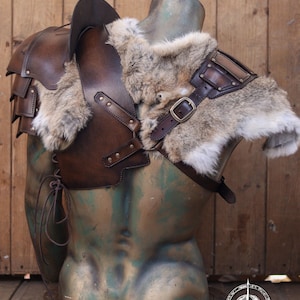 Mannequin bust wearing full barbarian leather shoulder armor set with crossed harness and riveted pauldrons. The armor is aged brown, handcrafted in vegetable-tanned leather with visible patina and hand-stitched details. Worn over a dark medieval shirt, this larp armor set includes sword scabbard on the back. Perfect for viking costume, post-apocalyptic larp, or warrior cosplay outfit.