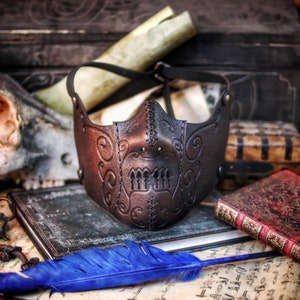 May include: A black leather mask with intricate stitching and a skull-like design. The mask is lying on a table with old books and a blue feather.