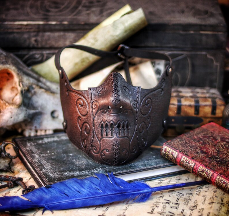 May include: A black leather mask with intricate stitching and a skull-like design. The mask is lying on a table with old books and a blue feather.