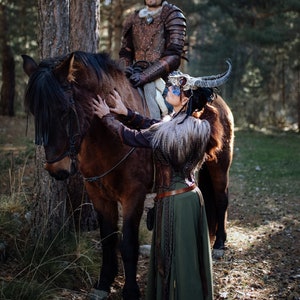 May include: A woman in a green dress and a horned headdress is petting a brown horse. A man in armor is sitting on the horse behind her.