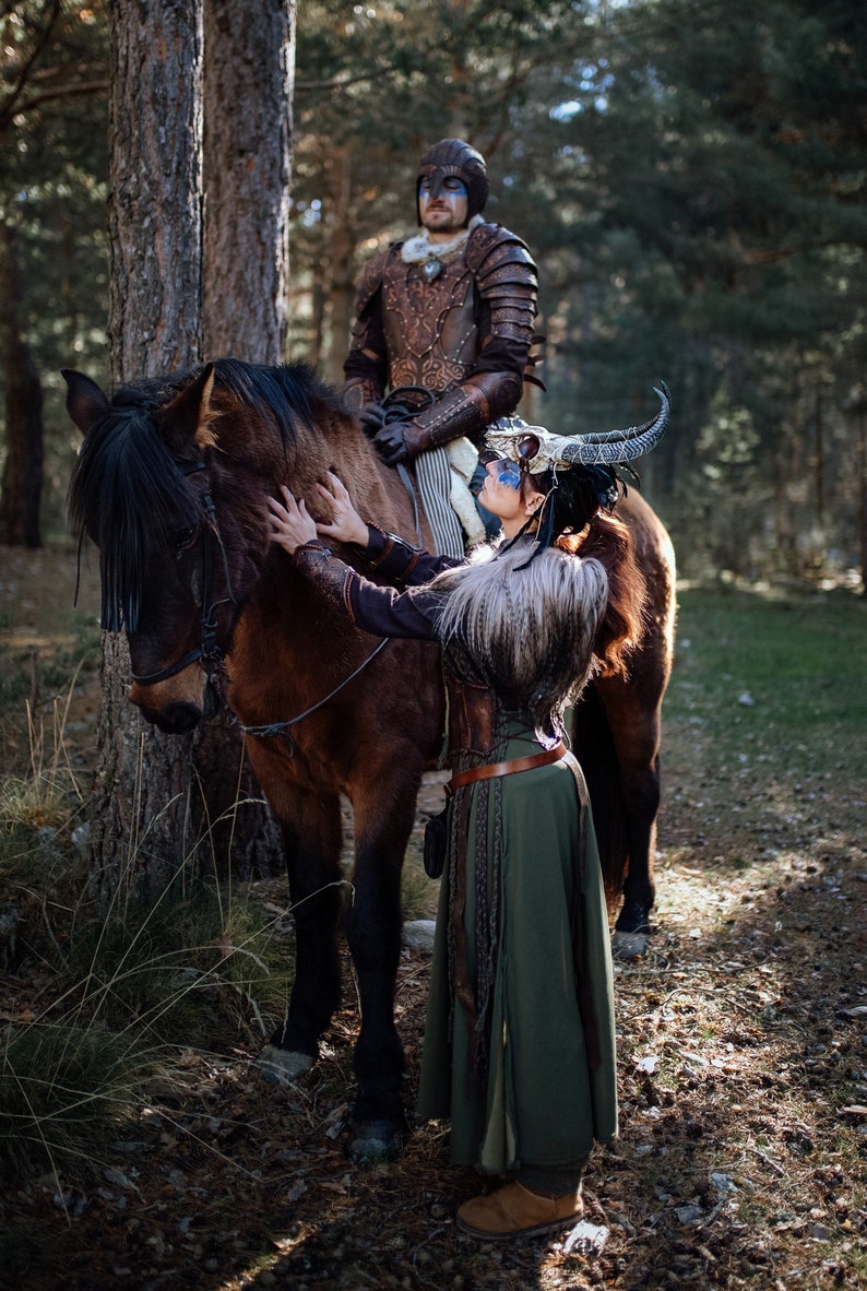 May include: A woman in a green dress and a horned headdress is petting a brown horse. A man in armor is sitting on the horse behind her.