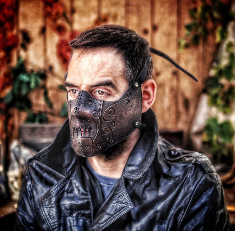A person wearing a black leather jacket and a brown leather mask with intricate designs. The mask covers the person's nose and mouth and has a stitched design around the edges. The mask has a skull-like design with a small, red, stitched mouth. Death Wizard Leather Mask M1 - Handcrafted Mask for Dark mages witches and powerful sorcerers with Intricate Details and Metallic Finish
