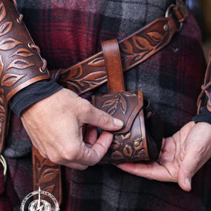 Close view of a person fastening a brown floral embossed leather coin pouch to a matching belt. The handcrafted botanical details and brass rivets stand out against the plaid skirt. The suede body expands to hold coins, dice or small items. Ideal for renaissance faire or renfaire costumes, this fantasy pouch fits elven characters, witches, herbalists and nobles seeking a functional and elegant leather accessory for LARP or cosplay events.