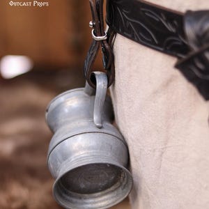 May include: A small, silver-colored metal jug with a handle, hanging from a dark brown leather strap attached to a black belt. The belt has a decorative embossed pattern. The text "Outcast Props" is visible in the upper left corner.