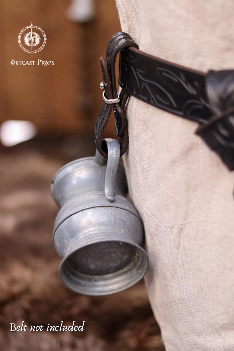May include: A small, silver-colored metal jug with a handle, hanging from a dark brown leather strap attached to a black belt. The belt has a decorative embossed pattern. The text "Outcast Props" is visible in the upper left corner.