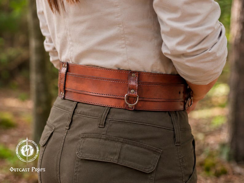 May include: A wide, brown leather belt featuring multiple horizontal stitched layers, metallic rivets, and integrated metal D-rings for attachment. The belt is worn over a light-colored shirt and olive green trousers, displaying a rugged, functional design.