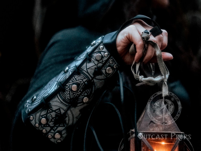 Female hand holding a lantern while wearing silver rune bracer with gothic engravings. Scene evokes mystery and magic, ideal for LARP witches, dark sorceresses, or fantasy rogues. Made with thick leather and aged painting technique, this handcrafted bracer offers full mobility, symbolic power and visual storytelling for immersive fantasy costumes.