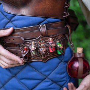 Könnte beinhalten: Ein brauner Ledergürtel mit dekorativen Haltern für kleine Glasflaschen. Die Flaschen enthalten farbige Flüssigkeiten. Eine größere, runde Flasche mit einem Korken ist ebenfalls sichtbar. Der Gürtel wird über einem blauen, gesteppten Kleidungsstück getragen.