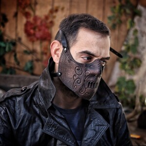 May include: A brown leather face mask with intricate designs and a skull-like pattern. The mask has a strap that goes around the head.