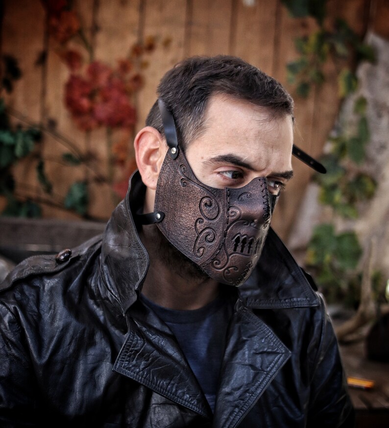 May include: A brown leather face mask with intricate designs and a skull-like pattern. The mask has a strap that goes around the head.