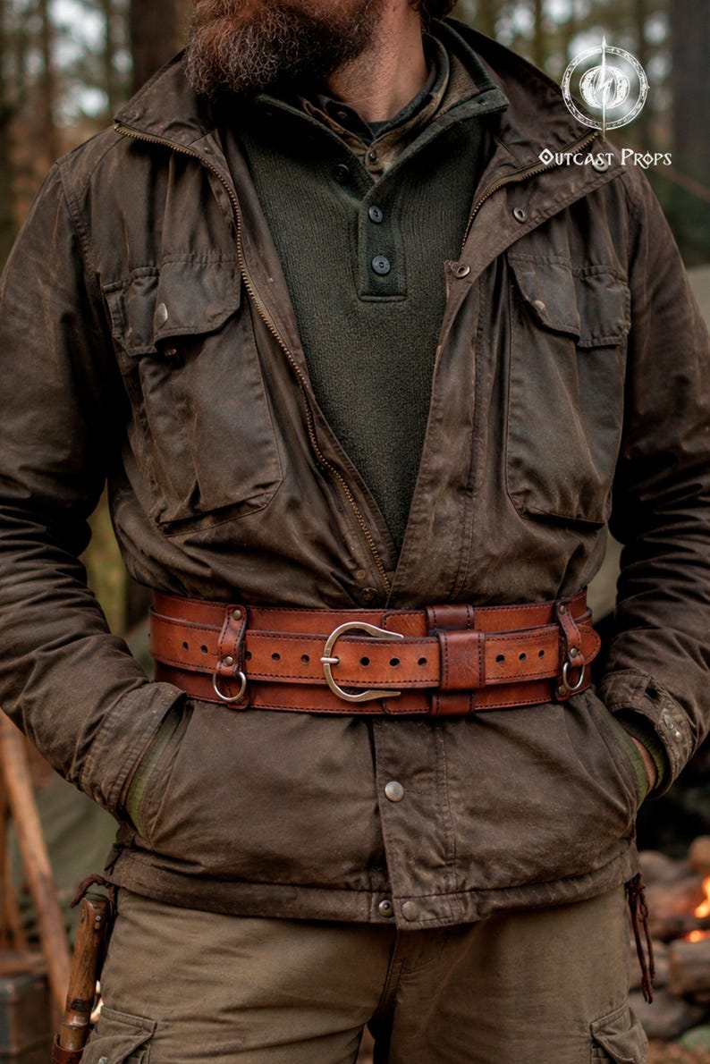 Aged brown bushcraft leather belt worn over an outdoor jacket in a forest camp setting, showing the belt as part of a rugged bushcraft and post apocalyptic outfit. Handmade wide utility belt with brass hardware, adjustable inner strap and side attachment rings for modular gear. Suitable for hiking gear, camping gear, tactical costume, military inspired outfit, steampunk belt, LARP armor, medieval ranger costume and Renaissance Faire clothing.