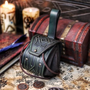 Green leather coin pouch resting on a fantasy map with scattered metal coins, books and candles in the background. The pouch features angular geometric embossing, brass rivets, suede sides and a sturdy leather belt loop. Made from vegetable-tanned leather and hand aged, it evokes Celtic, Viking and dwarf aesthetics. Designed for practical use during LARP, medieval fairs and cosplay, this leather coin pouch securely carries coins while adding a handcrafted, immersive detail