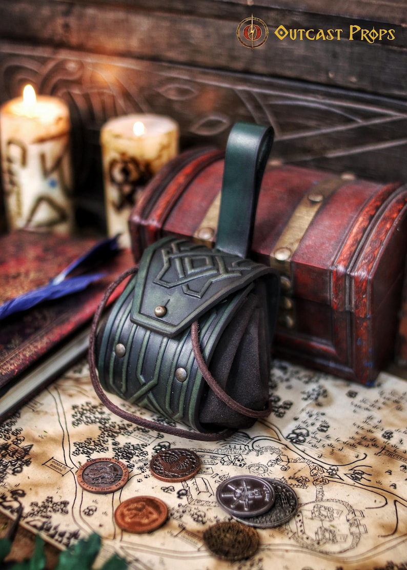 Green leather coin pouch resting on a fantasy map with scattered metal coins, books and candles in the background. The pouch features angular geometric embossing, brass rivets, suede sides and a sturdy leather belt loop. Made from vegetable-tanned leather and hand aged, it evokes Celtic, Viking and dwarf aesthetics. Designed for practical use during LARP, medieval fairs and cosplay, this leather coin pouch securely carries coins while adding a handcrafted, immersive detail
