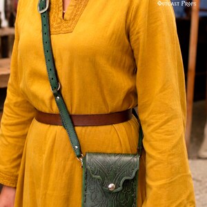 以下が含まれることがあります： 複雑なエンボス加工のフローラルパターン、シルバーメタルのスナップ留め、シルバーのバックルが付いた調節可能なグリーンのレザーストラップが特徴のダークグリーンのレザー製クロスボディバッグ。刺繍が施された長袖のマスタードイエローのチュニックの上に着用。