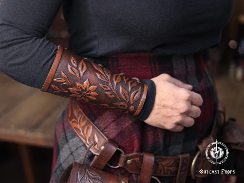 Side view of an aged brown floral leather bracer worn over a dark sleeve, paired with a renaissance faire belt bearing the same embossed leaf motif. The floral arm guard features hand tooled petals and natural shading that emphasize the artisan quality. This larp bracer functions as a fantasy cosplay accessory suited for elves, herbalists or fae characters, blending beautifully into medieval inspired outfits. Warm lighting highlights the engraved botanical pattern. Fae costume, ren faire, druid
