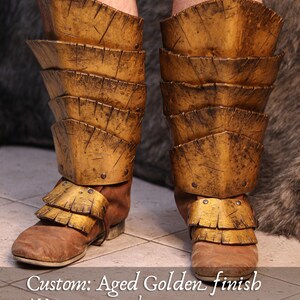 May include: Aged golden finish custom leg armor. The armor is made of layered plates with a distressed finish. The boots are brown leather. The text on the image says "Custom: Aged Golden finish".