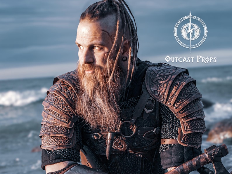 Bearded LARP warrior in full viking leather shoulder armor Sedrik, seated by the sea at sunset. The engraved Nordic pauldrons with layered leather plates feature detailed Celtic knots and fold for full mobility. Ideal for viking cosplay shoulder armor, Nordic warrior LARP gear, celtic leather pauldrons for fantasy costumes or medieval shoulder armor for immersive roleplay and conventions