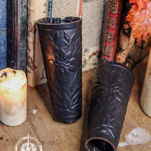 Two black floral leather bracers stand near antique books and warm candlelight in a medieval styled scene. The renaissance faire bracer design displays embossed leaves and petals, making each piece suitable as an elf cosplay accessory or fae costume accessory. The floral arm guard texture and artisan finish create a handcrafted larp bracer look ideal for fantasy or woodland themed characters. Fae costume, ren faire, druid larp, leaf design, floral design.