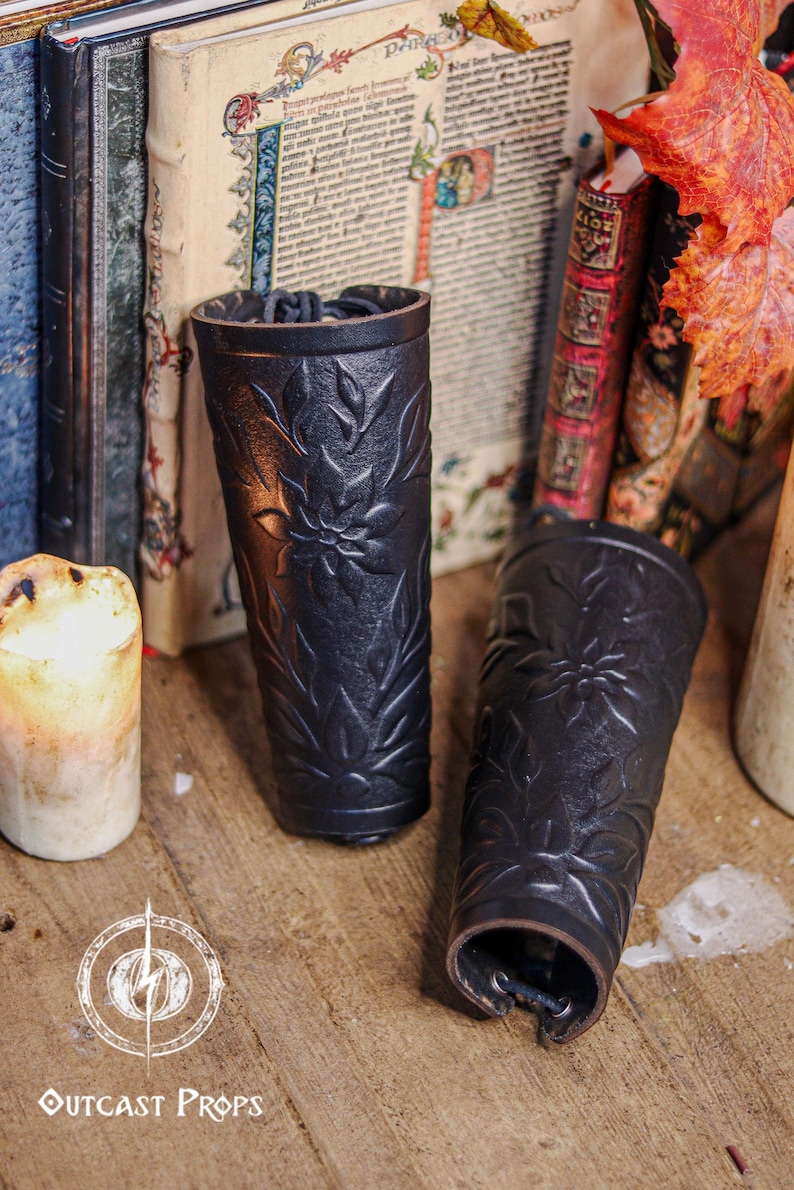 Two black floral leather bracers stand near antique books and warm candlelight in a medieval styled scene. The renaissance faire bracer design displays embossed leaves and petals, making each piece suitable as an elf cosplay accessory or fae costume accessory. The floral arm guard texture and artisan finish create a handcrafted larp bracer look ideal for fantasy or woodland themed characters. Fae costume, ren faire, druid larp, leaf design, floral design.