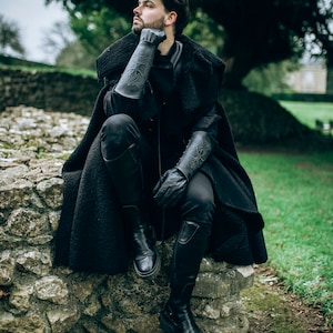 Fantasy adventurer wearing black leather and cloak sits on stone ruins, gazing into the distance. His arms feature long black leather bracers with Yggdrasil engraving, inspired by Norse mythology. This piece suits witcher armor, viking costume woman, or dark rogue cosplay. Perfect for LARP, ren faire, or medieval outfits, the bracers enhance any warrior or ranger look. The scenery evokes solitude and deep roleplay immersion in outdoor events.