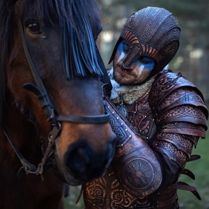 May include: A person wearing a leather helmet and armor with intricate designs, standing next to a brown horse with a blue fringe on its head.