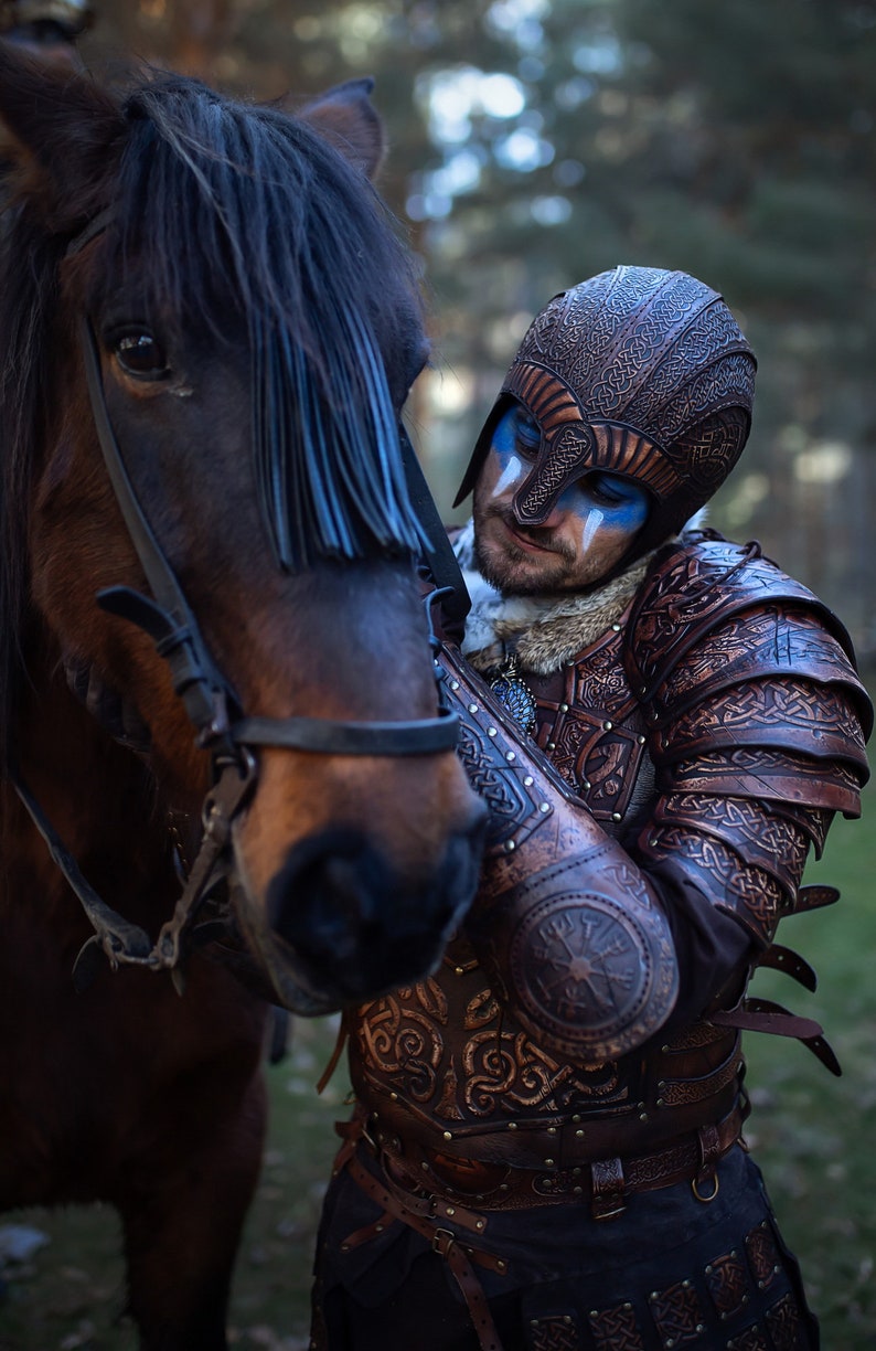 May include: A person wearing a leather helmet and armor with intricate designs, standing next to a brown horse with a blue fringe on its head.