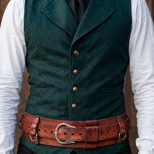 May include: A dark green vest with a subtle tonal paisley pattern, featuring four brass-toned buttons and a notched lapel. It is worn over a white long-sleeved shirt with a black cravat and a wide, brown leather belt with a silver-toned buckle.
