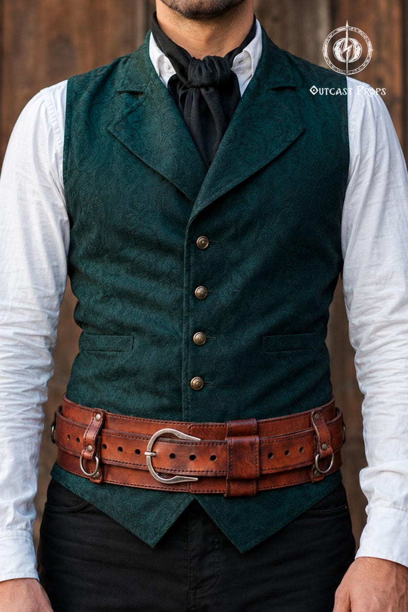 May include: A dark green vest with a subtle tonal paisley pattern, featuring four brass-toned buttons and a notched lapel. It is worn over a white long-sleeved shirt with a black cravat and a wide, brown leather belt with a silver-toned buckle.