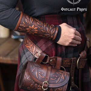 A close view of an aged brown floral leather bracer worn on the forearm, paired with a matching renaissance faire belt and floral pouch. The embossed petals and curling leaves create a detailed floral arm guard with warm shading ideal for fantasy outfits. The larp bracer sits over dark sleeves and plaid fabric, showing its handcrafted texture and suitability as an elf cosplay accessory or woodland character piece. Perfect for renaissance faire costumes and immersive roleplay looks. Fae druid