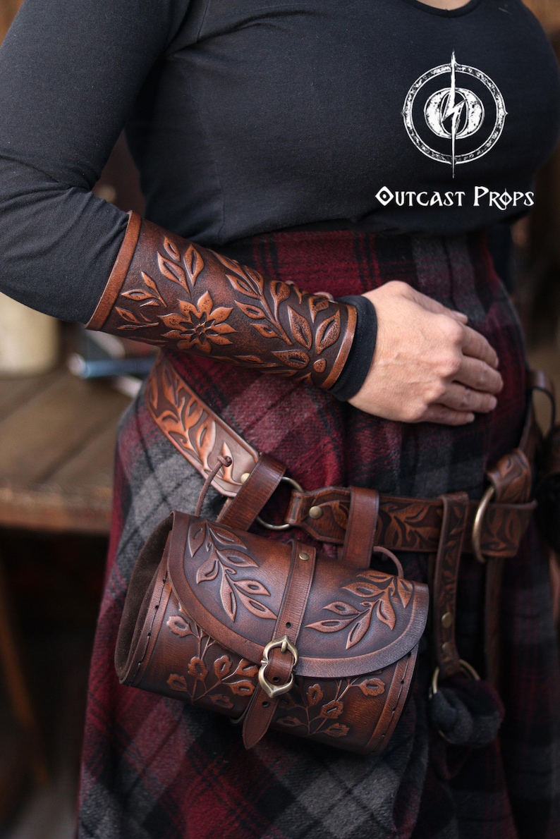 A close view of an aged brown floral leather bracer worn on the forearm, paired with a matching renaissance faire belt and floral pouch. The embossed petals and curling leaves create a detailed floral arm guard with warm shading ideal for fantasy outfits. The larp bracer sits over dark sleeves and plaid fabric, showing its handcrafted texture and suitability as an elf cosplay accessory or woodland character piece. Perfect for renaissance faire costumes and immersive roleplay looks. Fae druid