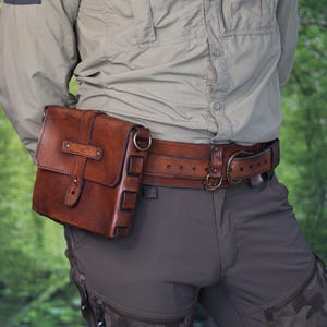 May include: A brown leather pouch with a flap closure, a central strap, and metal hardware. The pouch features side loops and is attached to a wide, matching brown leather belt with a metal buckle. The text Outcast Props is visible.