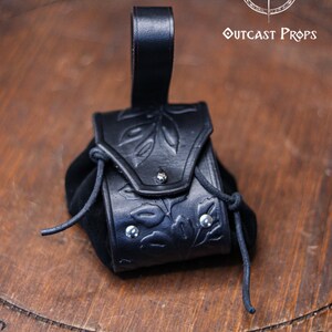A black leather coin pouch with embossed botanical motifs stands on a circular wooden board engraved with the Outcast Props logo. The pouch combines a structured vegetable tanned leather flap with a soft suede body and silver rivets. Dark and elegant, it works perfectly for witches, shadowy elves, nobles or herbalist characters at a renfaire or LARP event. This fantasy dice pouch attaches to the belt and holds coins or small items securely.