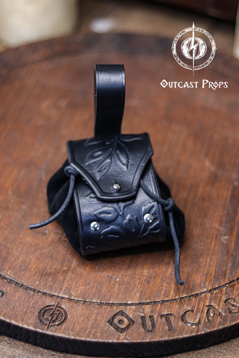 A black leather coin pouch with embossed botanical motifs stands on a circular wooden board engraved with the Outcast Props logo. The pouch combines a structured vegetable tanned leather flap with a soft suede body and silver rivets. Dark and elegant, it works perfectly for witches, shadowy elves, nobles or herbalist characters at a renfaire or LARP event. This fantasy dice pouch attaches to the belt and holds coins or small items securely.