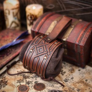 Decorated leather coin pouch with belt loop displayed beside a wooden chest and candles. Crafted from premium vegetable tanned leather with a hand-aged finish, embossed geometric motif, and suede body. Small medieval belt pouch for coins, dice, keys, jewelry, or tobacco. Fits fantasy, Viking, Celtic, dwarf aesthetics for LARP, Renaissance fairs, cosplay, or gifting.