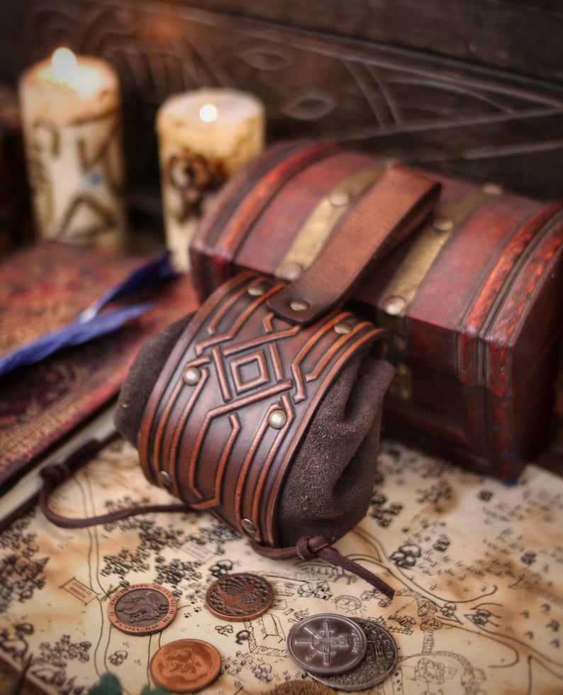 Decorated leather coin pouch with belt loop displayed beside a wooden chest and candles. Crafted from premium vegetable tanned leather with a hand-aged finish, embossed geometric motif, and suede body. Small medieval belt pouch for coins, dice, keys, jewelry, or tobacco. Fits fantasy, Viking, Celtic, dwarf aesthetics for LARP, Renaissance fairs, cosplay, or gifting.