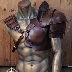 Mannequin bust wearing full barbarian leather shoulder armor set with crossed harness and riveted pauldrons. The armor is aged brown, handcrafted in vegetable-tanned leather with visible patina and hand-stitched details. Worn over a dark medieval shirt, this larp armor set includes sword scabbard on the back. Perfect for viking costume, post-apocalyptic larp, or warrior cosplay outfit.
