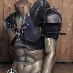 Pu&ograve; includere: Set di spallacci e imbracatura in pelle nera esposto su un manichino senza testa. L'armatura presenta una protezione per le spalle a strati, cinghie e fibbie metalliche. Il testo "SHOULDER PAULDRON + HARNESS" &egrave; in alto.