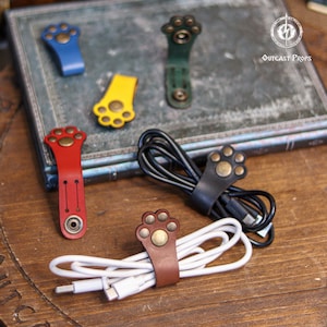 Puede incluir: Varios organizadores de cables de cuero con forma de huella de pata. Los organizadores son de varios colores: azul, amarillo, rojo, verde, marrón y negro. Cada organizador tiene un cierre de botón metálico. El logotipo de Outcast Props es visible.