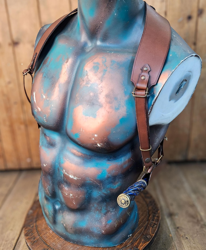 May include: A brown leather holster with adjustable straps for carrying a knife. The holster is worn on a mannequin with a blue and copper painted torso.