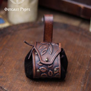A brown leather coin pouch with an embossed floral pattern stands centered on a round wooden board engraved with the Outcast Props logo. The soft suede sides create a rounded shape, while the looped strap allows it to hang from a belt. Warm brown tones and botanical carvings give it an elegant elven or herbalist look, ideal for witches, nobles and renfaire characters. Perfect as a leather coin bag or fantasy dice pouch for LARP, cosplay or daily use.