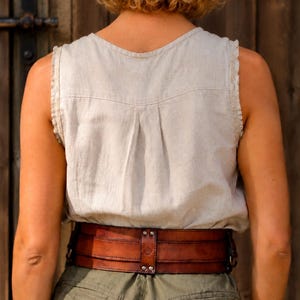 May include: A wide, dark brown leather corset belt featuring horizontal stitched panels, metal rivets, and a central vertical strap detail. The belt is worn over a light beige sleeveless linen top and olive green trousers, showcasing a structured design.
