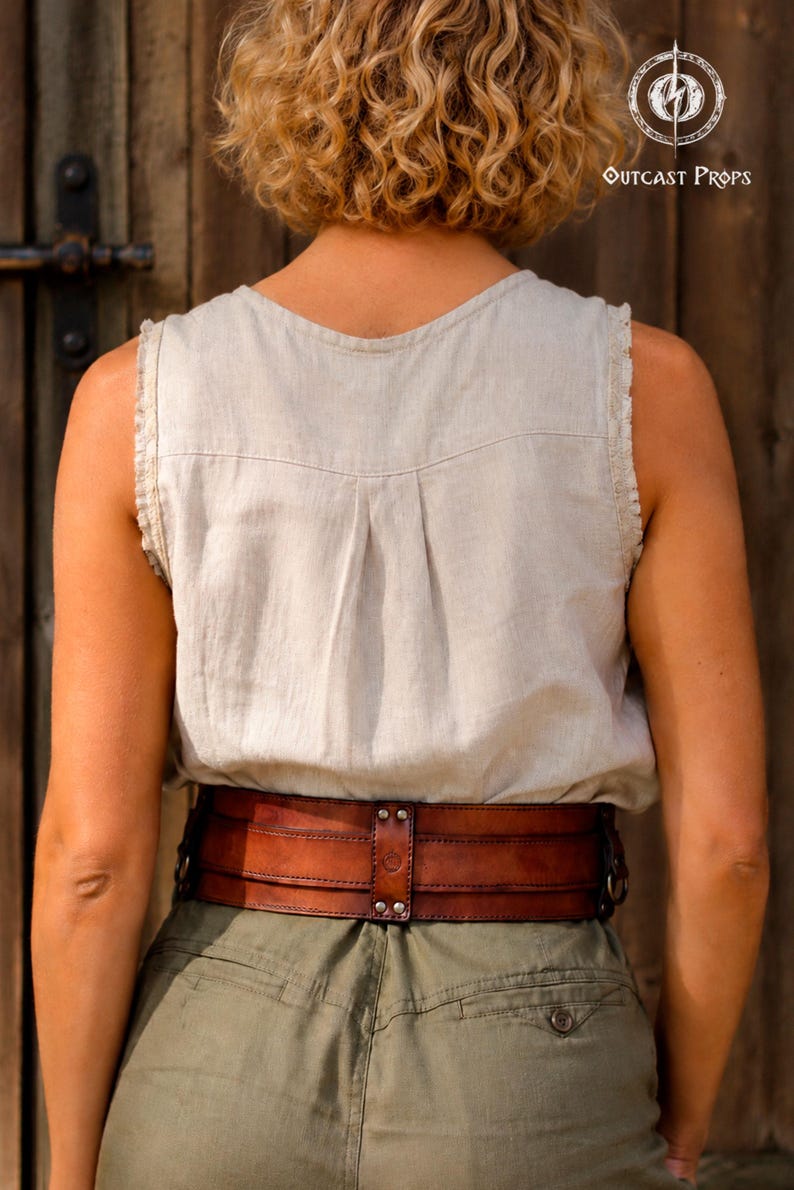 May include: A wide, dark brown leather corset belt featuring horizontal stitched panels, metal rivets, and a central vertical strap detail. The belt is worn over a light beige sleeveless linen top and olive green trousers, showcasing a structured design.