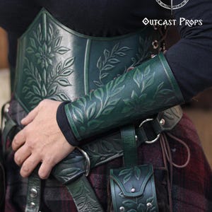 A floral leather bracer in aged green worn with a matching floral corset, shown in close up during a renaissance faire outfit. The renaissance faire bracer features embossed leaves and petals and works as an elf cosplay accessory, fae costume accessory or herbalist gear. Soft lighting highlights the engraved details and the elegant arm shaping, creating a refined larp bracer for fantasy costumes. Fae costume, ren faire, druid larp, leaf design, floral design.