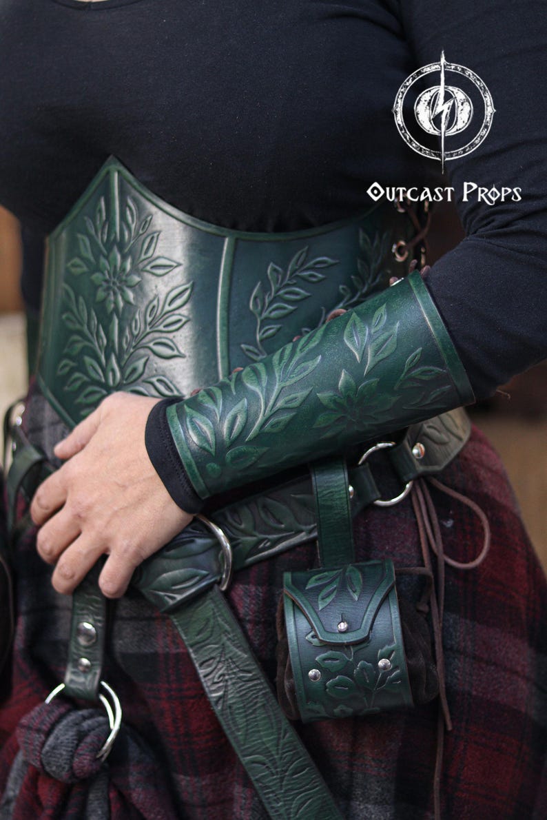A floral leather bracer in aged green worn with a matching floral corset, shown in close up during a renaissance faire outfit. The renaissance faire bracer features embossed leaves and petals and works as an elf cosplay accessory, fae costume accessory or herbalist gear. Soft lighting highlights the engraved details and the elegant arm shaping, creating a refined larp bracer for fantasy costumes. Fae costume, ren faire, druid larp, leaf design, floral design.