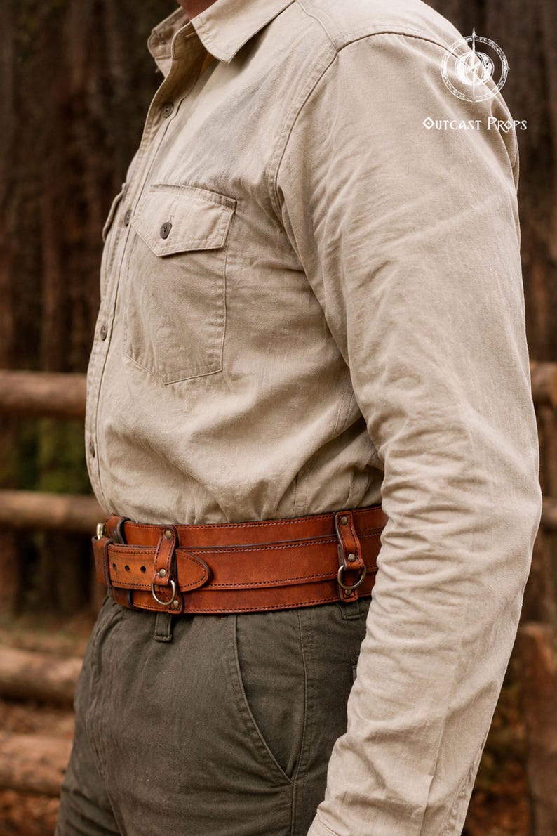 May include: A wide, tan leather belt featuring two horizontal straps, metal D-rings, and rivets. The belt is worn over dark olive trousers and a beige button-down shirt. The text "OUTCAST PROPS" is visible in the upper right corner of the image.