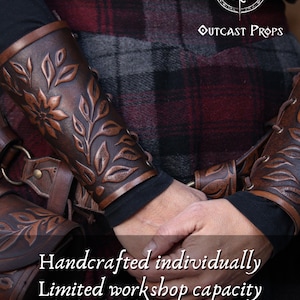 Two aged brown floral leather bracers are worn on crossed arms above a richly textured renaissance faire belt with a matching embossed pouch. The floral arm guard design features carved leaves and petals with deep highlights that emphasize the artisan finish. This larp bracer set fits naturally into medieval styled clothing, offering a fantasy cosplay accessory suitable for druids, elves or ranger characters. Warm browns and soft lighting enhance the handcrafted detail. Fae druid